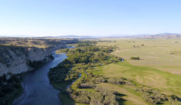 Aerial view of recreational land with a river winding through a green valley, rocky cliffs on one side, and open grassy plains stretching to distant mountains under a clear blue sky. Ideal hunting property with stunning natural scenery.