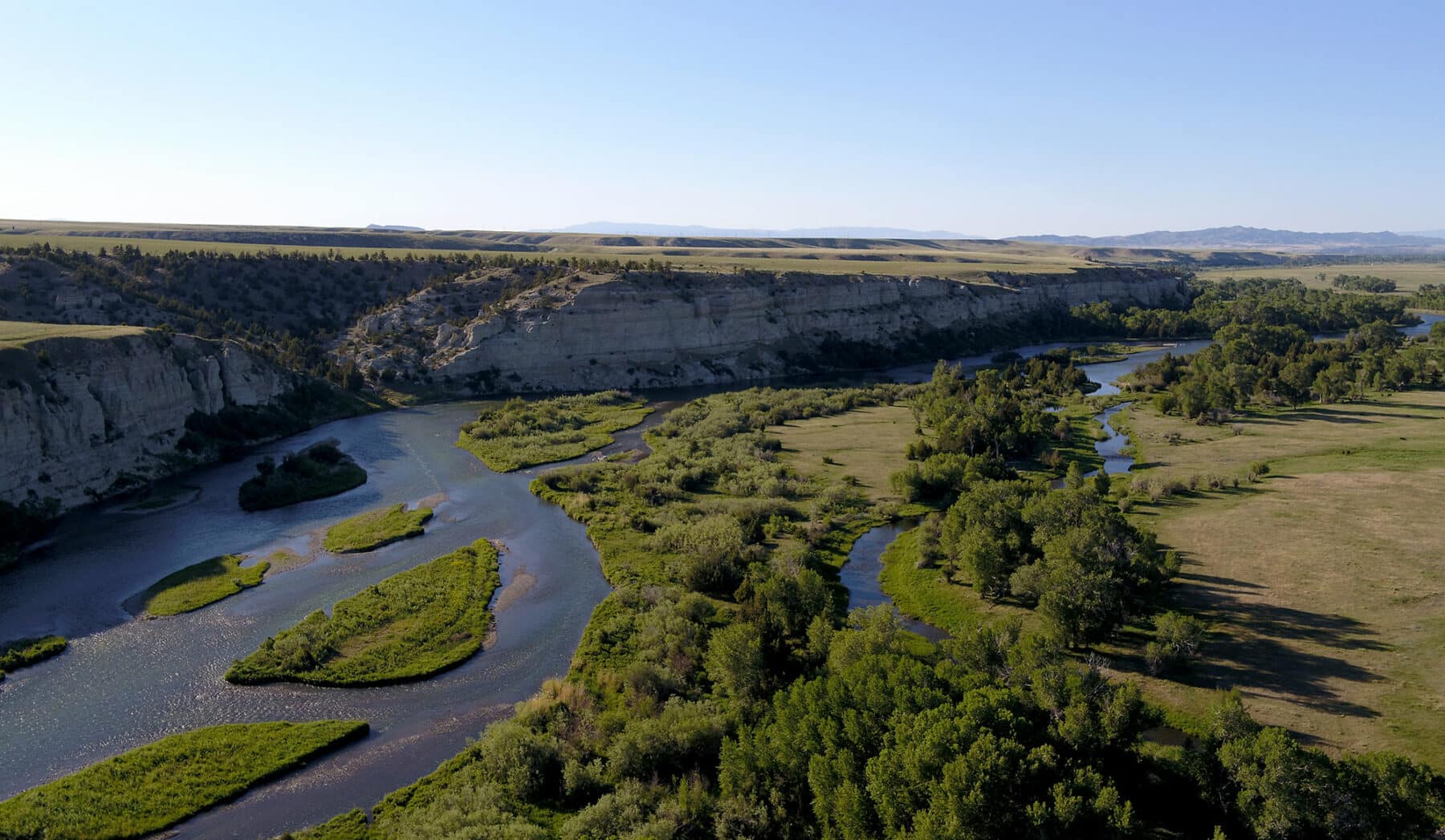 A wide river winds through a green, tree-lined valley with rocky cliffs and flat grasslands under a clear blue sky—ideal recreational land or potential cattle ranch.