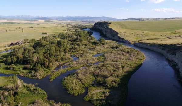 A winding river flows through a green, open landscape with clusters of trees, grassy fields, and distant mountains under a blue sky. A cattle ranch is visible near the riverbank on the left side of the image.