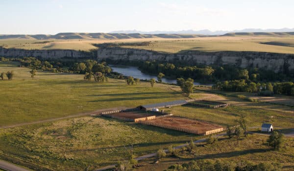 Aerial view of a rural landscape featuring a fenced cattle ranch with a barn and small white building near a river, surrounded by green fields, trees, cliffs, and distant hills under a clear sky.