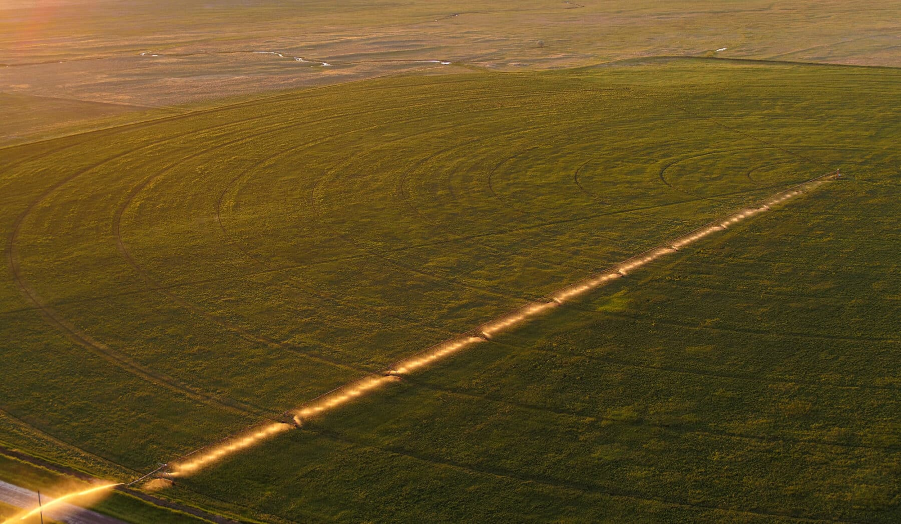 Aerial view of recreational land with circular irrigation patterns, bisected by a sunlit dirt path running diagonally across the green field at sunset.