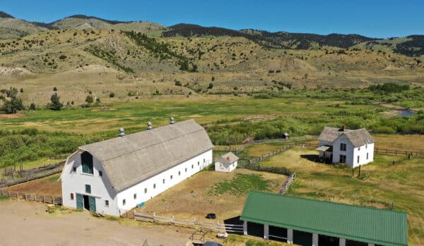 Aerial view of a rural farm and cattle ranch with a large white barn, white farmhouse, green-roofed shed, fenced areas, and rolling hills and mountains in the background under a clear blue sky; ideal land for sale.