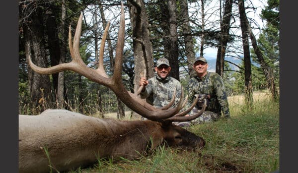 Two men in camouflage kneel in a forest beside a large elk with impressive antlers, smiling and holding its antlers. Surrounded by trees and natural light, this scene captures the adventure possible on your own ranch for sale.