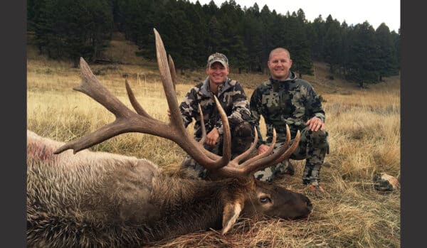 Two people in camouflage pose outdoors in a grassy field beside a large elk with antlers lying on the ground, showcasing the possibilities of this hunting property surrounded by pine trees and rolling hills.