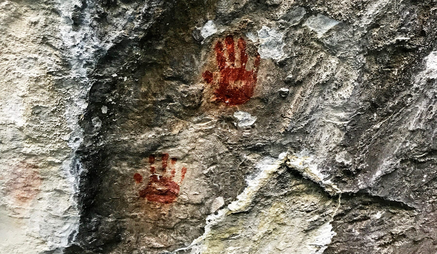 Two red handprints are painted on a rough, gray stone surface, resembling ancient cave art. The wall’s textures and natural cracks evoke the history found on a traditional cattle ranch or hidden corners of land for sale.