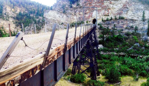 A rusty, elevated railroad bridge with wooden and metal supports leads into a dark tunnel in a rocky cliff, surrounded by green trees and rugged terrain—perfect for a hunting property or ranch for sale.