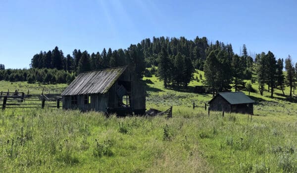 An old, weathered wooden barn and a small shed stand in a grassy field, surrounded by a split-rail fence, with forested hills and clear blue sky—ideal recreational land or hunting property for sale.