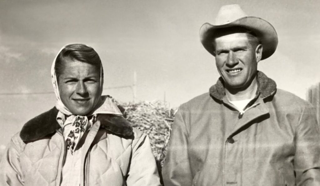 A black-and-white photo of a woman in a headscarf and quilted jacket stands beside a man in a cowboy hat and heavy coat outdoors, both squinting at the camera on what appears to be a cattle ranch.