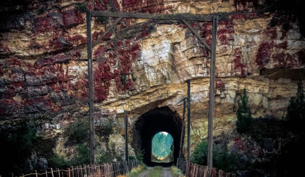 A dirt path winds through a rock tunnel cut into a cliff with red and yellow layers, framed by old wooden beams and wire fencing. The tunnel opens to a sunlit forest, highlighting the beauty of this recreational land or potential cattle ranch.