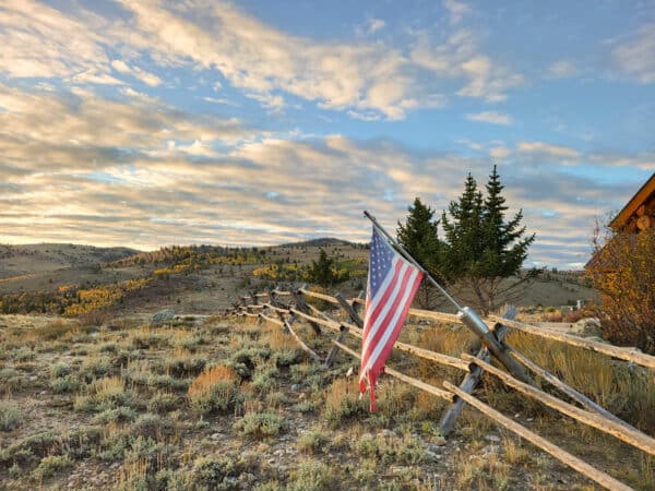 An American flag hangs on a rustic wooden fence in a grassy, hilly landscape with scattered trees and a partly cloudy sunset sky. A wooden cabin is partially visible to the right—ideal ranch for sale or hunting property seekers.