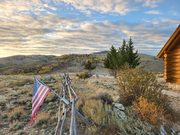 A rural landscape with hills and scattered trees under a partly cloudy sky. An American flag hangs from a wooden fence near a log cabin with autumn foliage, ideal as recreational land or for those seeking a scenic cattle ranch.