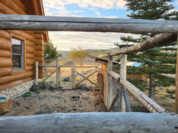 View through a rustic wooden fence toward a gate, with a log cabin to the left and trees, rocks, and a sunlit landscape in the background—ideal for recreational land or as a hunting property.