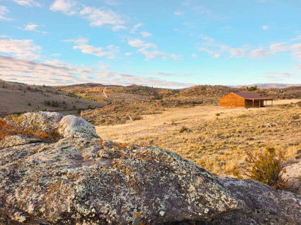 A rustic wooden shed stands on a sunlit, grassy hillside of recreational land surrounded by rolling, rocky terrain under a partly cloudy blue sky with distant hills in the background.