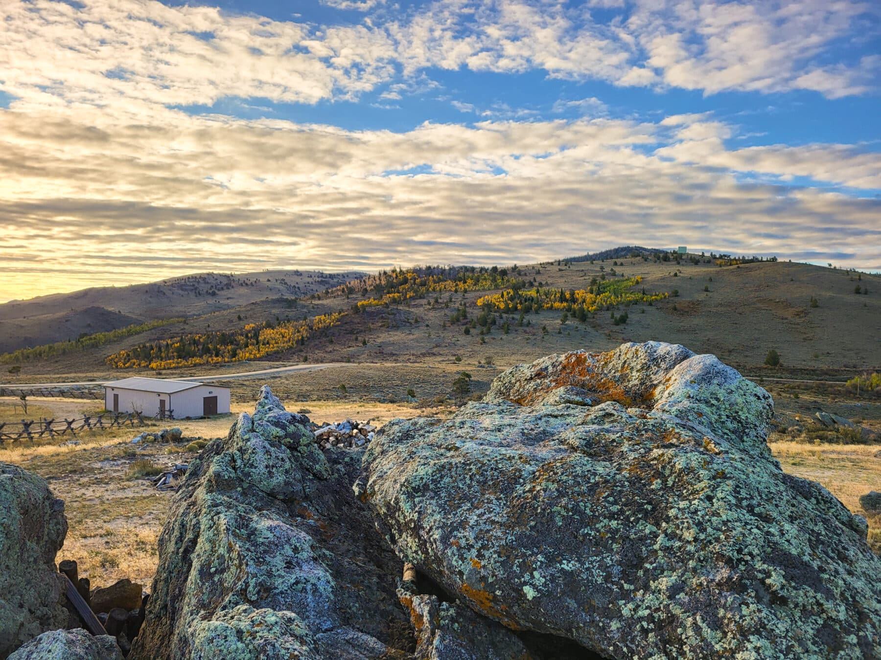 Large moss-covered rocks in the foreground overlook recreational land with rolling hills, yellow-leaved trees, a small building, and a dramatic sky filled with scattered clouds.