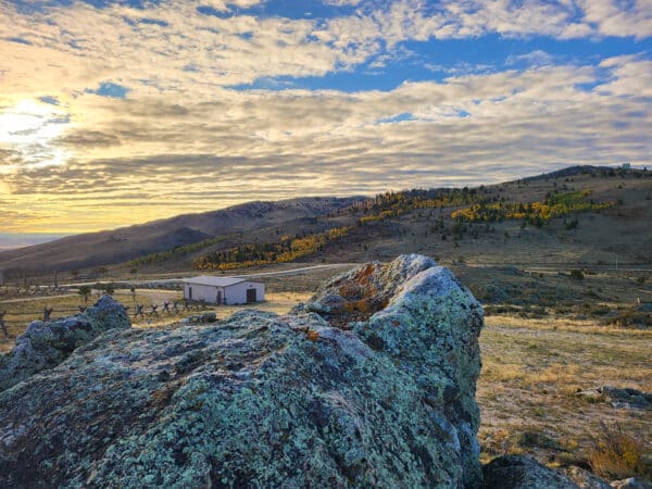 A rocky foreground with moss-covered boulders overlooks a small white building on open grassland, perfect recreational land for sale, with rolling hills, scattered trees, and a partly cloudy sky lit by the setting sun.