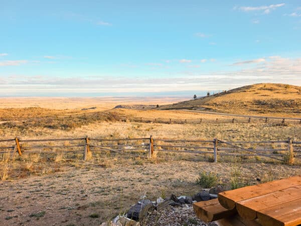 A wooden fence lines a dry, grassy landscape with rolling hills under a blue sky. In the foreground, part of a wooden picnic table is visible, highlighting recreational land ideal for relaxing in warm, early morning or late afternoon light.