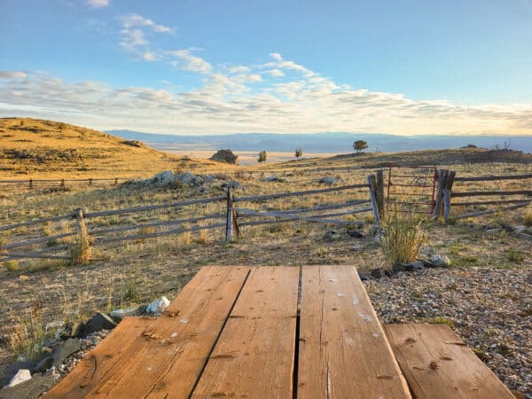 A wooden picnic table sits on rocky ground near a rustic split-rail fence, overlooking an open field with rolling hills—ideal land for sale perfect for a future cattle ranch under a blue sky with scattered clouds.