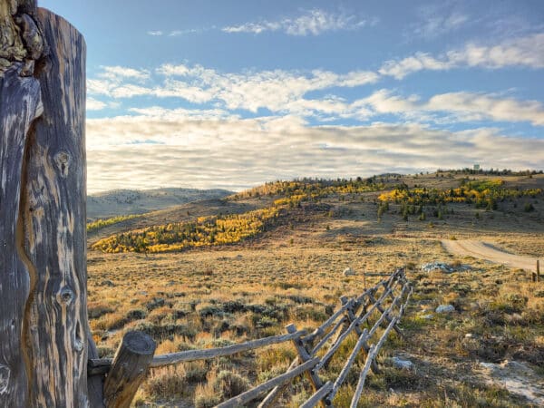 A rustic wooden fence runs through a dry, grassy field with rolling hills covered in yellow autumn trees under a blue sky with scattered clouds. Sunlight gently illuminates this scenic recreational land, ideal for a ranch for sale.