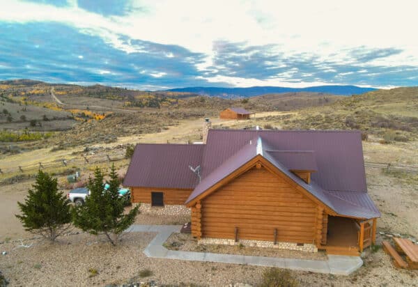 A wooden log cabin with a metal roof sits in a rural, hilly landscape with scattered trees under a cloudy sky. A white vehicle is parked beside the house, perfect for those seeking a peaceful ranch for sale amid distant hills stretching to the horizon.