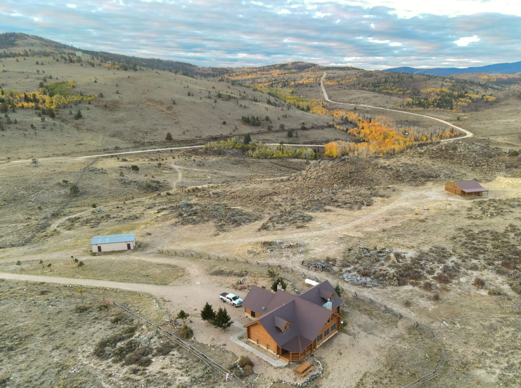 Aerial view of a wooden cabin and two outbuildings on recreational land in a dry, hilly landscape with scattered trees and dirt roads. Autumn foliage appears in the distance under a partly cloudy sky.