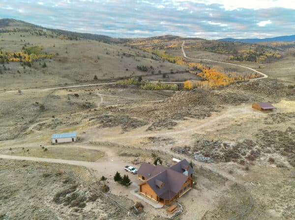 Aerial view of a wooden cabin and two outbuildings on recreational land in a dry, hilly landscape with scattered trees and dirt roads. Autumn foliage appears in the distance under a partly cloudy sky.