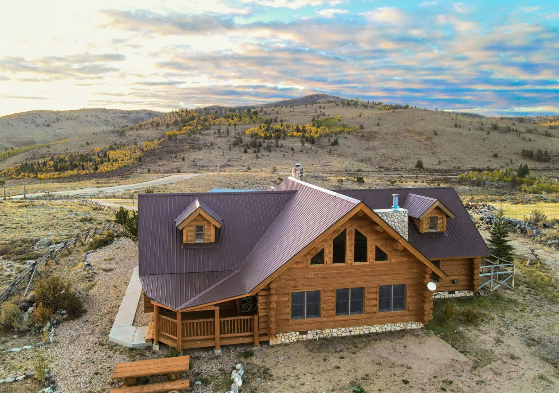 A rustic log cabin with a metal roof sits on recreational land in an open, hilly landscape under a partly cloudy sky, surrounded by sparse trees and dry grass, with rolling hills in the background.