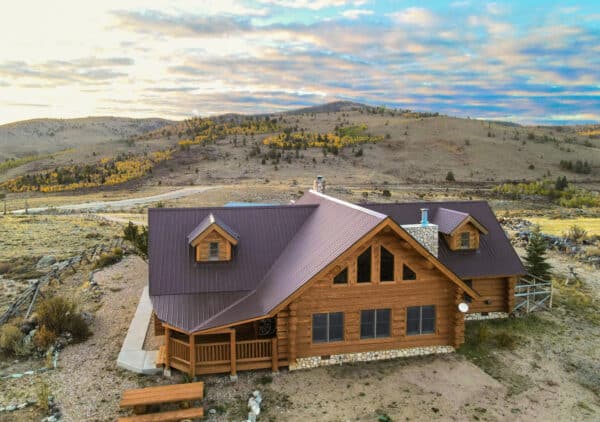 A rustic log cabin with a metal roof sits on recreational land in an open, hilly landscape under a partly cloudy sky, surrounded by sparse trees and dry grass, with rolling hills in the background.