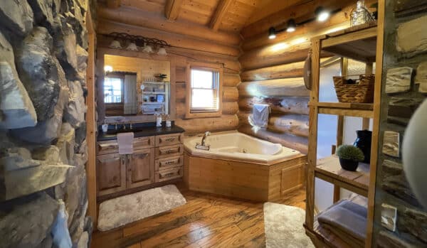 A cozy log cabin bathroom with wooden walls, a corner bathtub, twin sinks with a large mirror, open shelves with towels and decor, and soft lighting—perfect for relaxing after a day on your hunting property or recreational land.