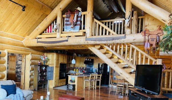 Cozy log cabin interior with wooden walls, exposed beams, and a loft area. The kitchen features rustic cabinets, Western decor, and saddles—perfect for a cattle ranch or recreational land. A staircase leads to the upper level with a TV in the foreground.