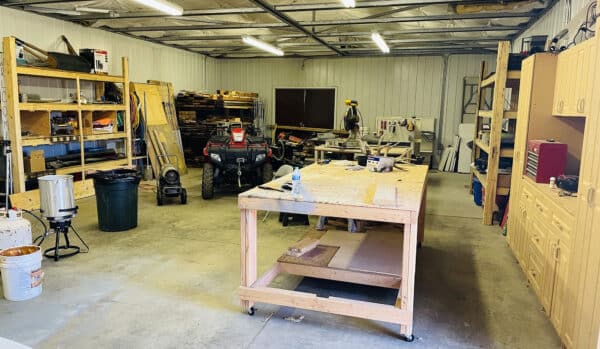 A spacious workshop with wooden shelves, tools, a center workbench, an ATV, and equipment neatly organized along the walls. The bright space is ideal for maintaining a cattle ranch or preparing recreational land. Overhead fluorescent lights ensure clear visibility.
