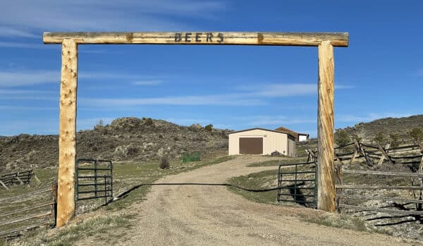 A wide wooden ranch gate labeled BEERS frames a gravel driveway leading to a beige barn with a brown roof, set against rocky hills under a clear blue sky. This scenic cattle ranch features fence lines along both sides of the driveway.