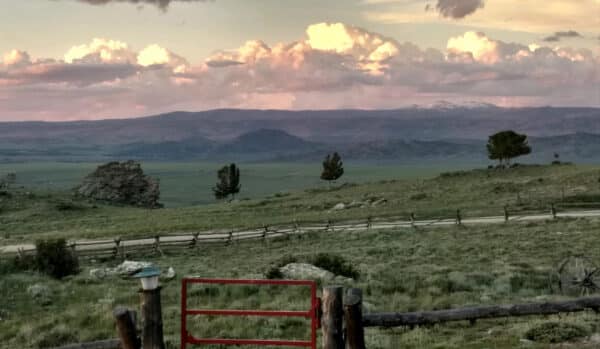 A scenic view of a grassy plain with wooden fences and scattered trees on a cattle ranch. A red gate sits in the foreground, with distant hills and mountains beneath dramatic sunset clouds.