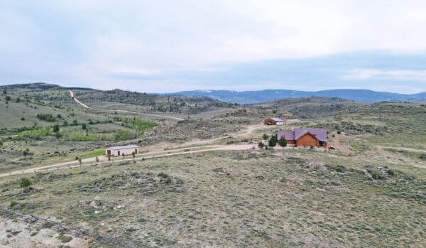 Aerial view of a rural landscape featuring a wooden house, smaller buildings, dirt roads, sparse trees, rolling hills, and mountains under a cloudy sky—ideal as recreational land or a hunting property.