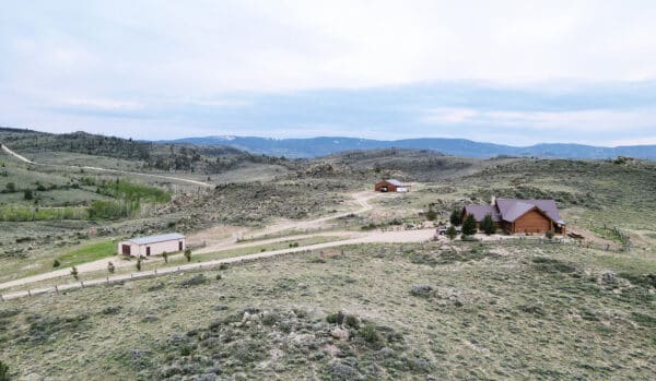 Aerial view of recreational land featuring a large wooden house, barn, and outbuildings, surrounded by rolling hills, scattered trees, and distant mountains under a cloudy sky.