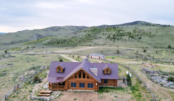 Aerial view of a wooden log cabin with a red metal roof, surrounded by a fenced yard on a cattle ranch, set in a grassy, hilly landscape with mountains in the background and a dirt road nearby.