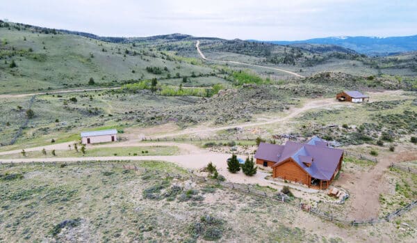 Aerial view of a wooden house and two outbuildings on a large, open hilly landscape with sparse vegetation, winding dirt roads, and green hills—perfect as a hunting property or recreational land—set against distant mountains under a cloudy sky.