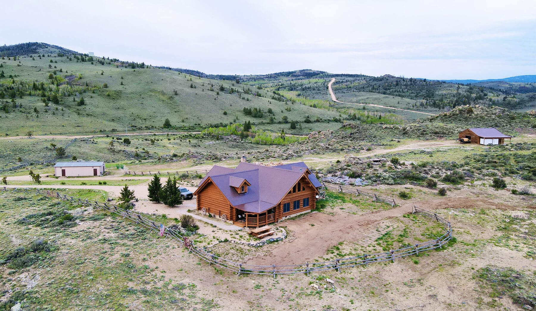Aerial view of a wooden cabin with a fenced yard in a hilly rural landscape, ideal as hunting property or recreational land, surrounded by sparse vegetation and mountains, with two outbuildings and a winding road nearby.