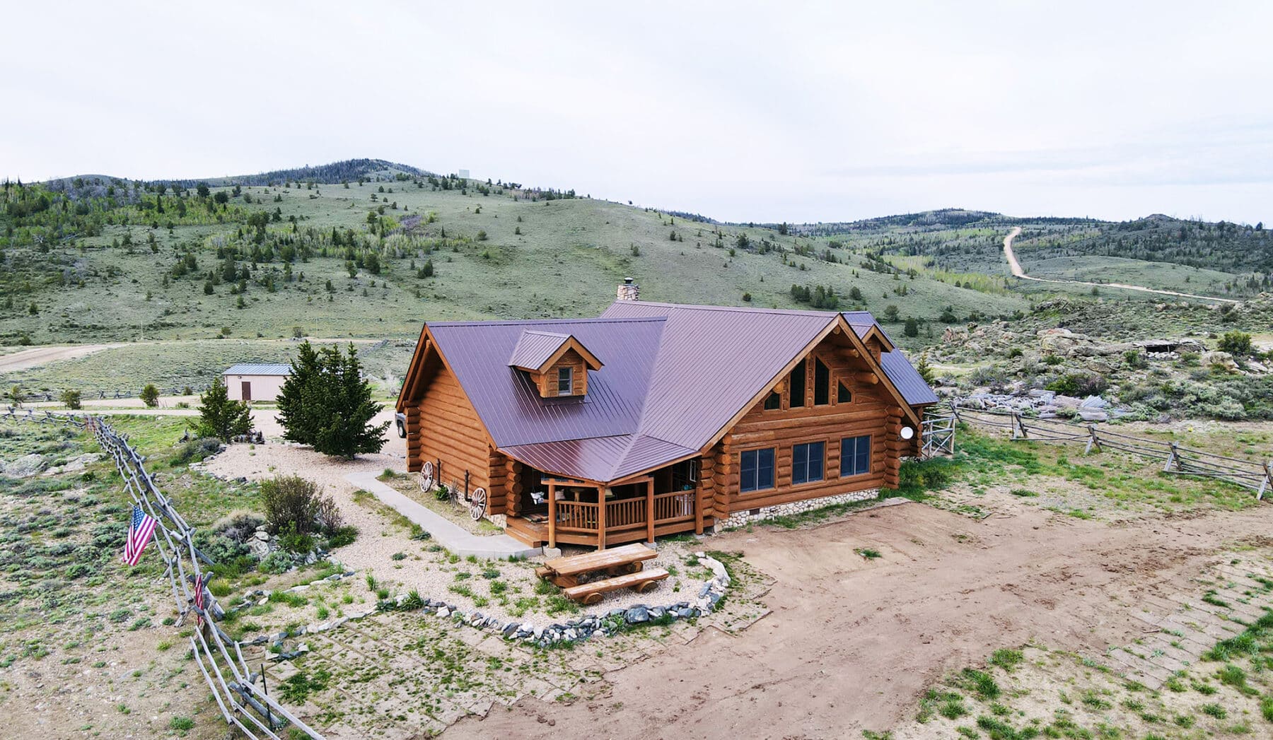 A wooden cabin with a metal roof sits on a dirt plot in a rural, hilly landscape with sparse green vegetation and trees, surrounded by wooden fencing. This ranch for sale features a flag and gravel pathway near the entrance.