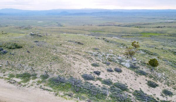 A wide, open landscape with grassy hills, scattered shrubs, a dirt road, and a barbed wire fence in the foreground—ideal recreational land or hunting property. Mountains are visible in the distance under a cloudy sky.