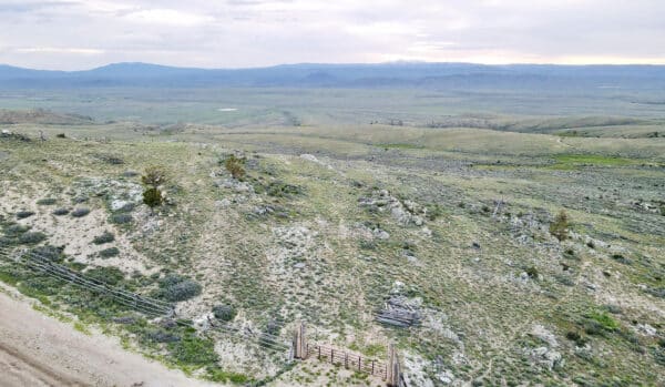 A wide, open landscape with rolling hills covered in sparse green vegetation, a wooden fence and gate in the foreground, and distant mountains under a cloudy sky—perfect land for sale ideal as a cattle ranch.