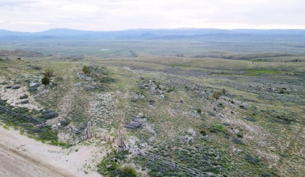 Aerial view of recreational land with rolling green hills, sparse vegetation, and distant mountains under a cloudy sky. An old wooden gate and fence line are visible near a dirt road in the foreground—ideal hunting property.