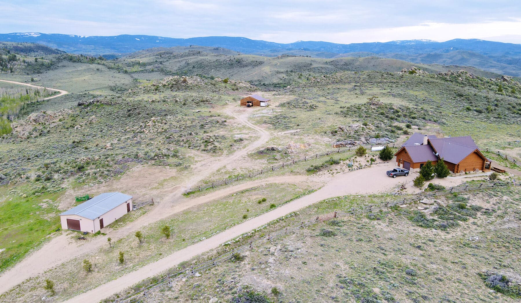 Aerial view of a rural landscape with a log cabin, two outbuildings, dirt roads, and rolling hills in the background under a cloudy sky—ideal as a ranch for sale or hunting property.