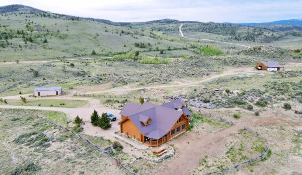 Aerial view of a log cabin house with a brown roof, surrounded by a dirt road, scattered trees, and rolling green hills. Two outbuildings sit nearby on this scenic ranch for sale in a rural, mountainous landscape.