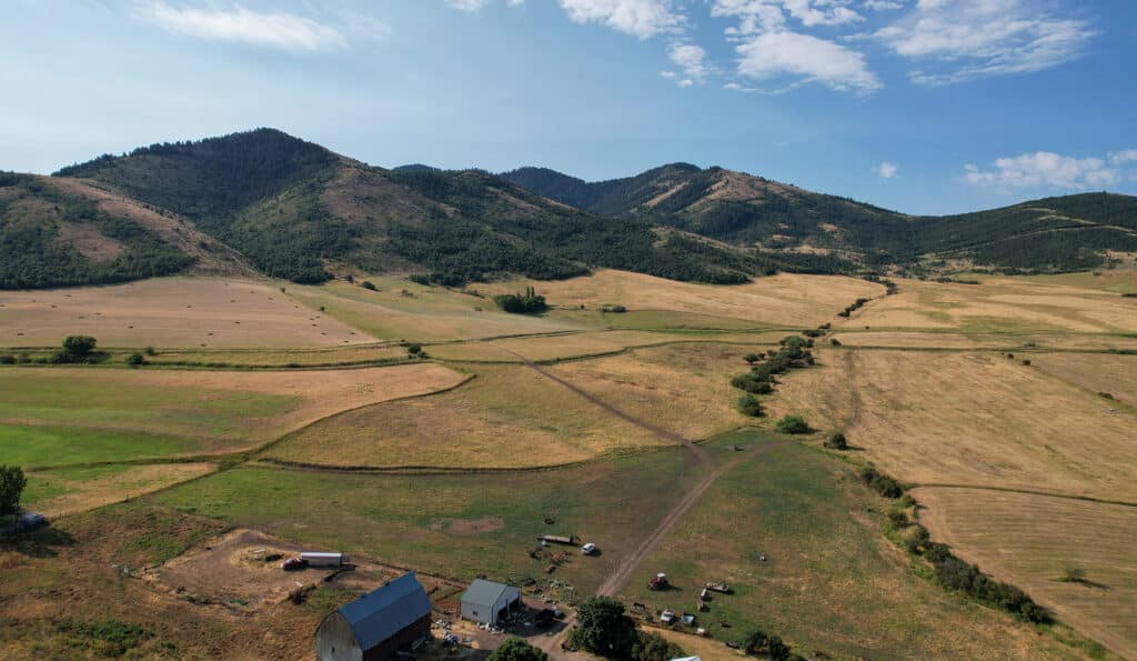 Aerial view of a rural landscape with rolling hills, fields of dry grass, scattered hay bales, and a few farm buildings under a partly cloudy sky—ideal as a cattle ranch or hunting property.