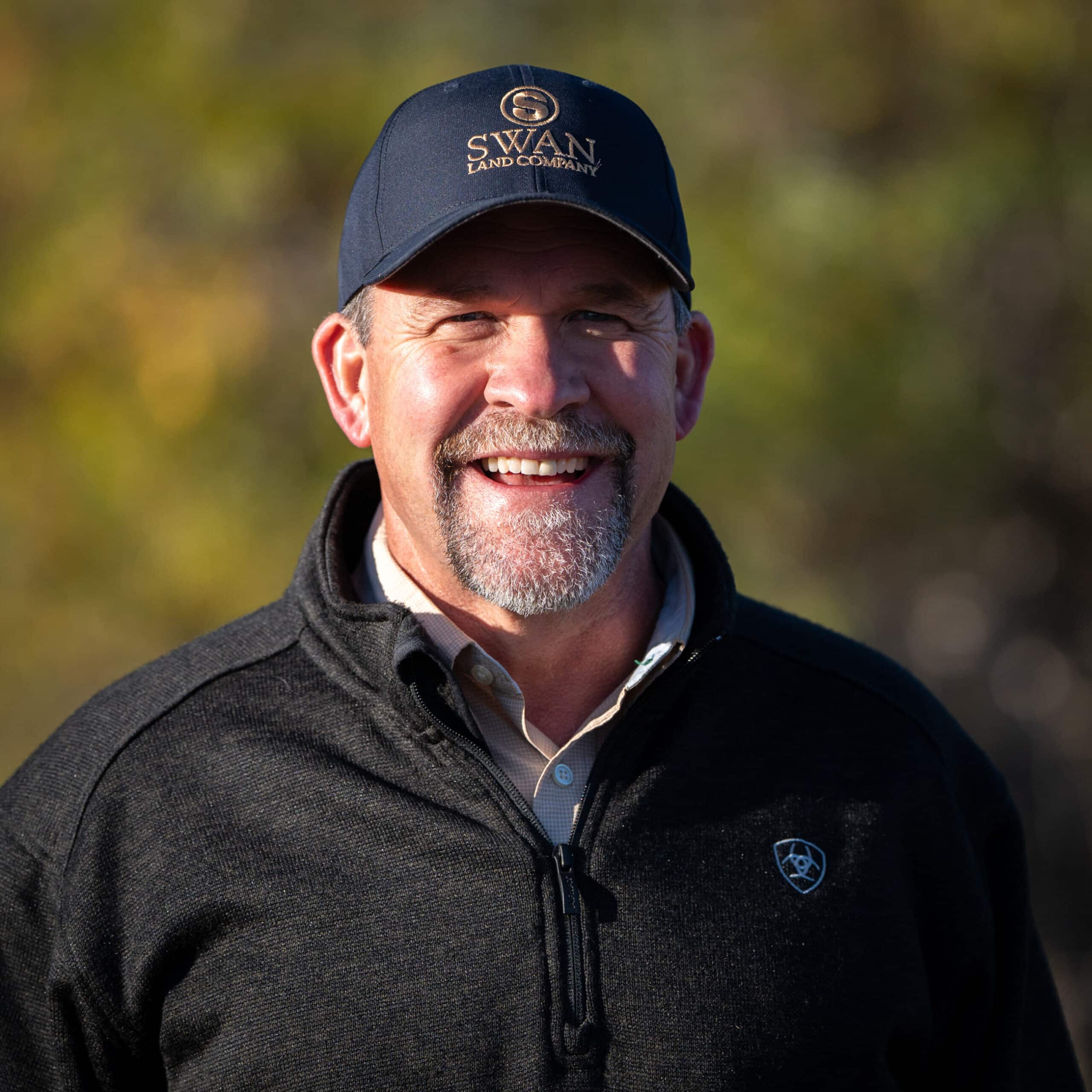 A smiling man wearing a navy cap with Swan Land Company logo and a dark zip-up jacket stands outdoors in natural sunlight, with a blurred green and yellow background—perfect for those interested in ranch for sale or hunting property.