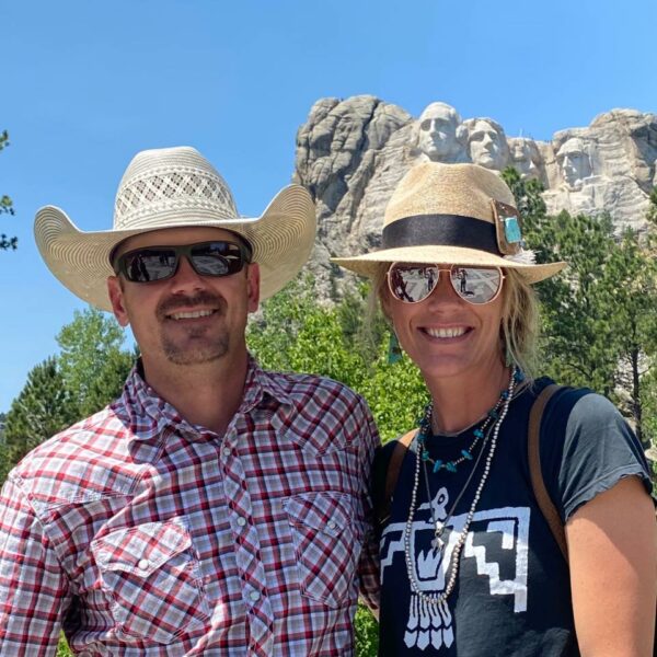 A smiling man and woman wearing hats and sunglasses pose outdoors with Mount Rushmore in the background, enjoying the clear blue sky—perfect for exploring nearby ranch for sale.
