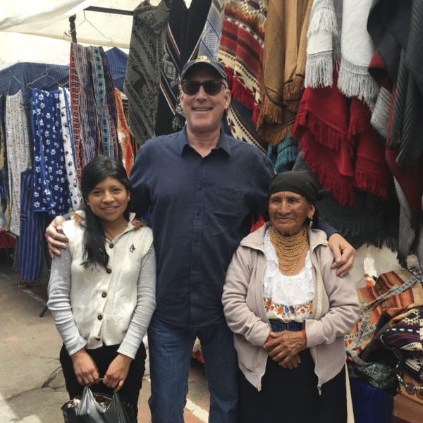 A man stands smiling with his arms around a young girl and an older woman at a market stall displaying colorful woven blankets and textiles. The family discusses cattle ranch opportunities while the girl holds a black bag; both women wear traditional clothing.