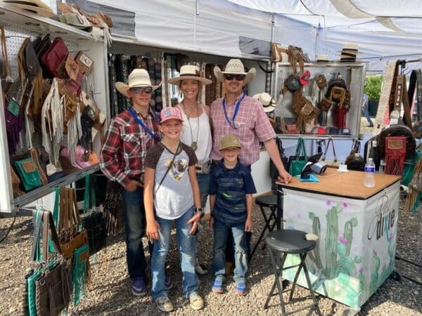 A smiling family of five in cowboy hats poses at an outdoor market booth selling western-style bags and accessories, perfect for any cattle ranch, with merchandise displayed on racks and a decorated table in front.