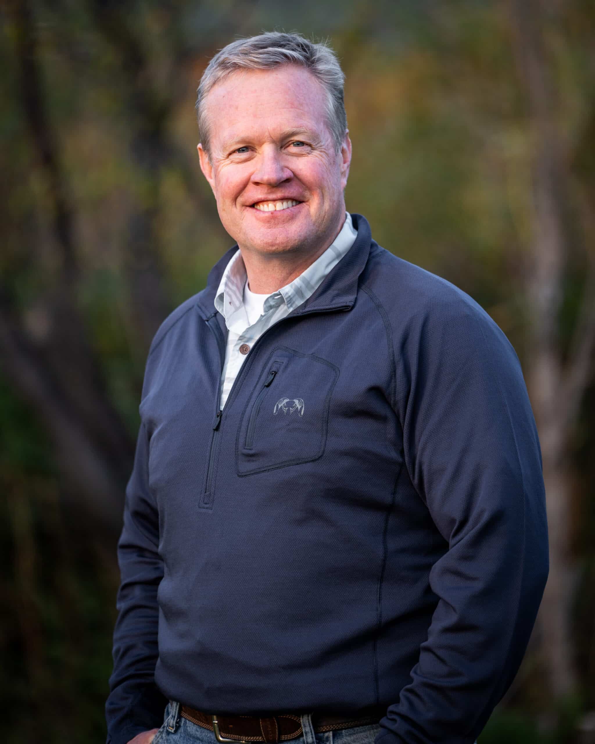A man with short blond hair, in a blue pullover and light gray collared shirt, stands outdoors smiling, with soft, blurred greenery in the background—ideal scenery for a cattle ranch or hunting property land for sale.
