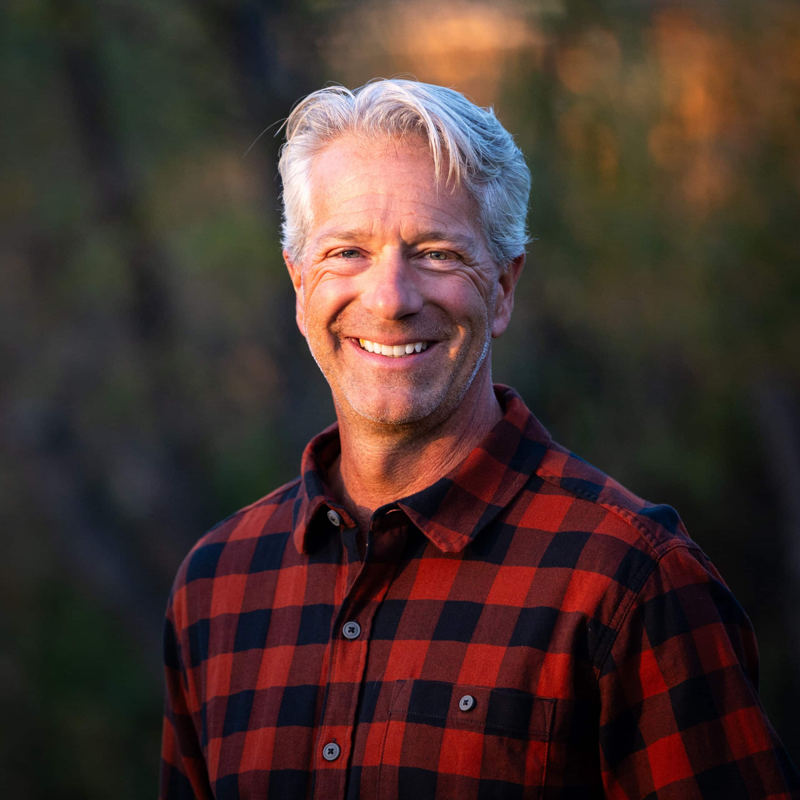 A smiling older man with short gray hair, wearing a red and black plaid shirt, stands outdoors on recreational land with a blurred green and brown background.